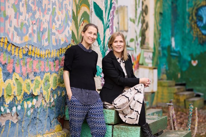 Kaksi henkilöä seisoo värikkään, koristeellisesti maalatun seinän edessä ulkona.