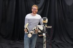 Quentin Houbre seisoo hymyillen, pukeutuneena raidalliseen paitaan ja farkkuihin, ja pitää kädessään robottirakennelmaa.