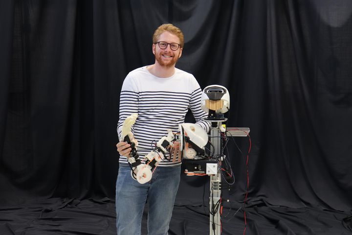 Quentin Houbre seisoo hymyillen, pukeutuneena raidalliseen paitaan ja farkkuihin, ja pitää kädessään robottirakennelmaa.