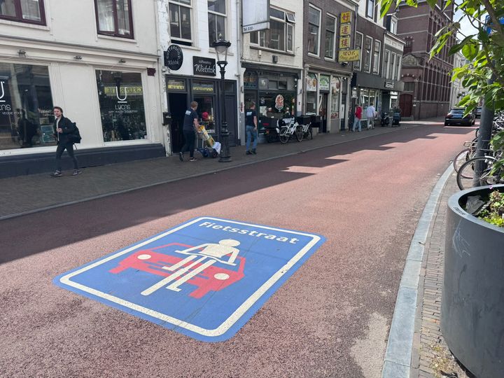 Pyöräkatu Utrechtissa, jossa on sininen pyöräkatusymboli ajoradalla. Kadun varrella on liikkeitä ja jalankulkijoita.