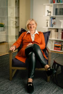 Marjo Miettinen istuu modernissa nojatuolissa, pukeutuneena oranssiin takkiin ja nahkahousuihin.