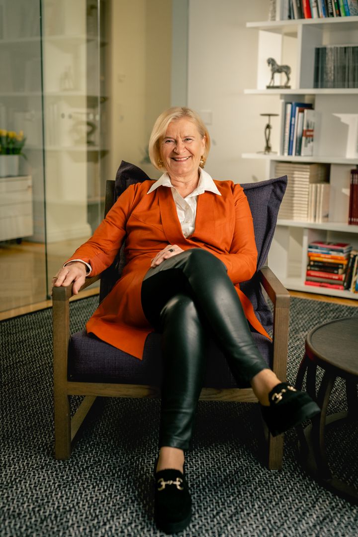 Marjo Miettinen istuu modernissa nojatuolissa, pukeutuneena oranssiin takkiin ja nahkahousuihin.