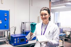 Nainen laboratoriotakissa esittelee pientä esinettä laboratoriossa, jossa on laitteita taustalla.