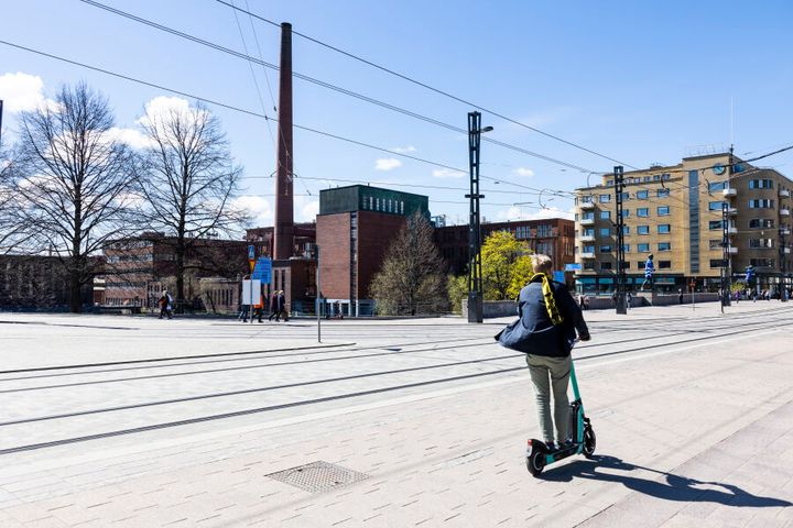Henkilö ajaa sähköpotkulaudalla kaupunkimaisemassa, taustalla rakennuksia ja sininen taivas.