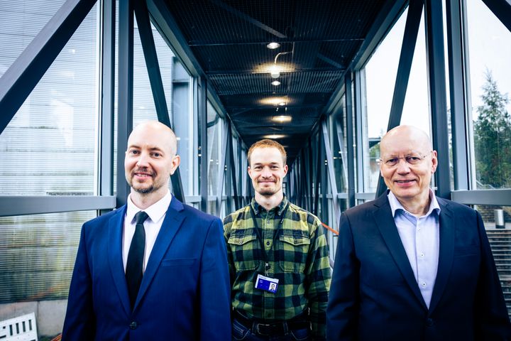 Kolme miestä seisoo toimiston käytävällä, joista yksi on puvussa ja muut rennommissa vaatteissa.