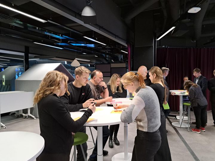Ryhmä ihmisiä keskustelemassa seisten korkeiden pöytien ääressä avarassa, teollishenkisessä tilassa, taustalla näkyy teknologialaitteita.