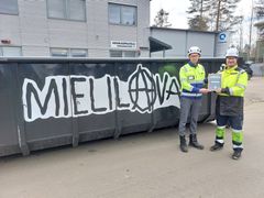Kaksi miestä työvaatteissa seisovat Kuusakosken Joensuun-toimipisteessä. Yksi heistä ojentaa kunniakirjan toiselle. Taustalla näkyy kierrätyslava, jossa lukee "MIELILAVA".