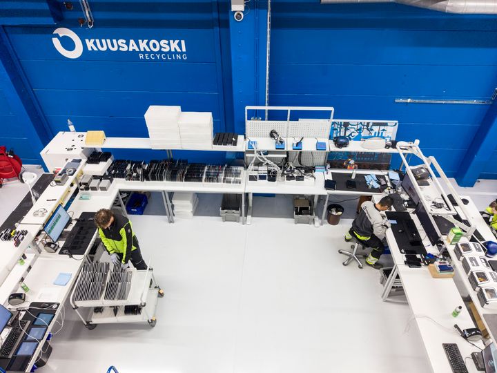 Lintuperspektiivikuva Kuusakoski Recyclingin tuotantotilasta, jossa työntekijät käsittelevät ja lajittelevat elektroniikkalaitteita pitkillä työpöydillä.