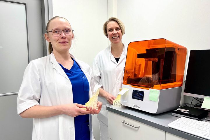 Erikoishammaslääkäri Helena Mehtonen ja radiologian erikoislääkäri Irina Rinta-Kiikka seisovat pöytätason vieressä. Pöydällä on 3D-tulostin, jossa on oranssi läpinäkyvä kansi.
