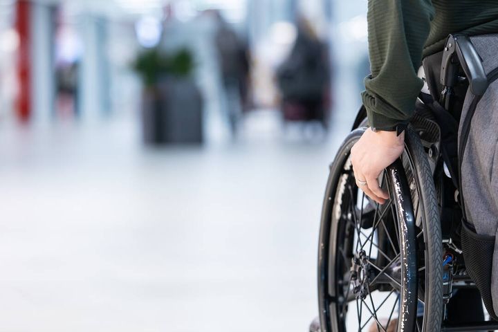 Kuvassa näkyy pyörätuolissa istuvan henkilön käsi ja pyörätuolin rengas. Taustalla on sumeana sairaalan käytävä.