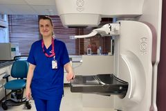 Annukka Salminen sinisissä työvaatteissaan seisoo mammografialaitteen vierellä ja hymyilee.