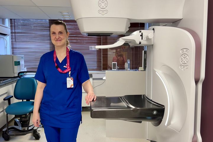 Annukka Salminen sinisissä työvaatteissaan seisoo mammografialaitteen vierellä ja hymyilee.