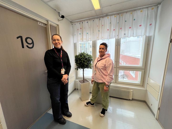 Kaksi naista seisoo käytävällä vastaanottohuoneen oven edessä, taustalla on ikkuna.