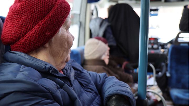 Ikääntynyt istuu bussin penkillä, ja takana näkyy muita matkustajia.