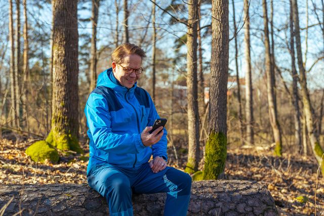 Mies on ulkona luonnossa, istuu kivellä ja käyttää kännykkää.