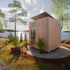Vaalea suuri-ikkunainen kuutiosauna seisoo terassilla veden äärellä, saunan katolla on aurinkopaneeli