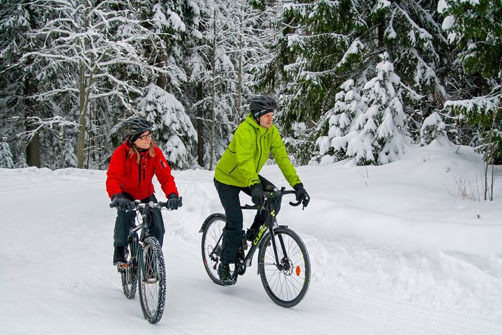 Kaksi aikuista pyöräilee lumisella tiellä talvimaisemassa.