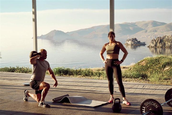 Kaksi henkilöä treenaamassa kuntosalilla meren äärellä. Toinen tekee askelkyykkyä painon kanssa, toinen seisoo kädet lanteilla.
