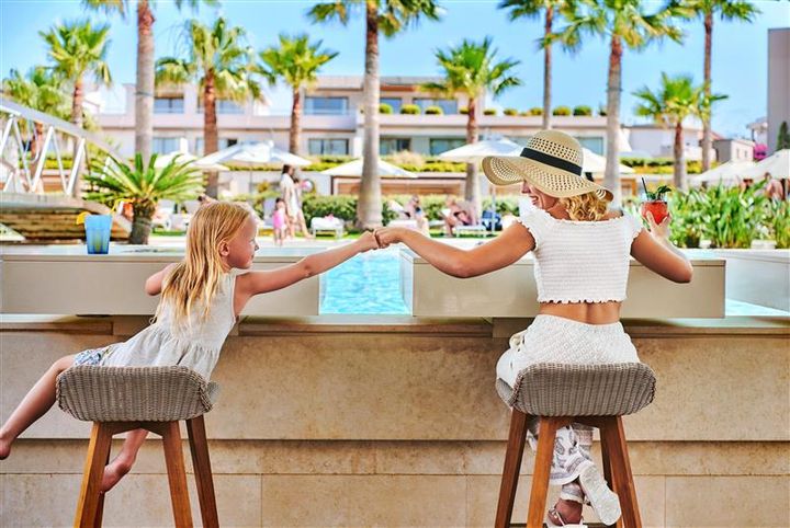 Äiti ja tytär istuvat baaritiskillä ulkona, taustalla uima-allas ja aurinkotuoleja.