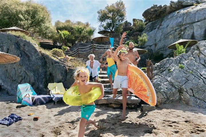 Ihmisiä rannalla, lapset juoksevat iloisina uimalelut kainalossa.