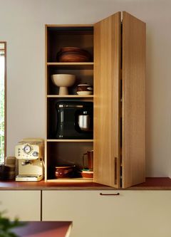 Valoisa keittiö, jossa on avonainen puinen kaappi taitto-ovilla. Kaapissa on kulhoja, vaahdotin ja espressokone.