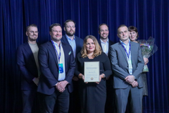 Ryhmä ihmisiä seisoo yhdessä; keskiössä henkilö, joka pitelee palkintoa.