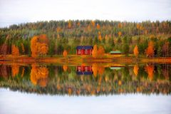 Punainen talo järven rannalla, ympärillä syksyinen metsä ruskan sävyissä.