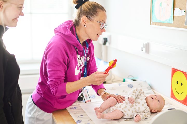 Nainen hoitaa vauvaa hoitopöydällä sairaalaympäristössä. Toinen henkilö seisoo vieressä.