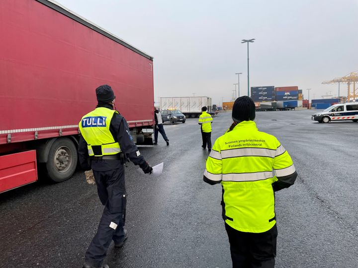 Sumuinen loppusyksyn päivä Vuosaaren satamassa Helsingissä. Vasemmalla näkyy kirkkaanpunainen rekan perävaunu. Sen vierellä, kuvan etualalla, kävelee kaksi henkilöä keltaisissa huomiovaatteissa. Toisella lukee takin selässä Tulli, toisen selässä Suomen ympäristökeskus.