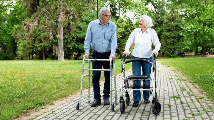 Äldre man och kvinna promenerar med rollatorer på en stig i en park.