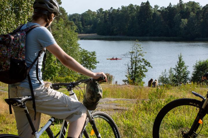 Valokuvassa on etualalla ihminen polkupyörän selässä katsomassa taka-alalla olevalle kalliorannalle, jossa istuu muutama ihminen. Vedessä on kaksi kanottia.