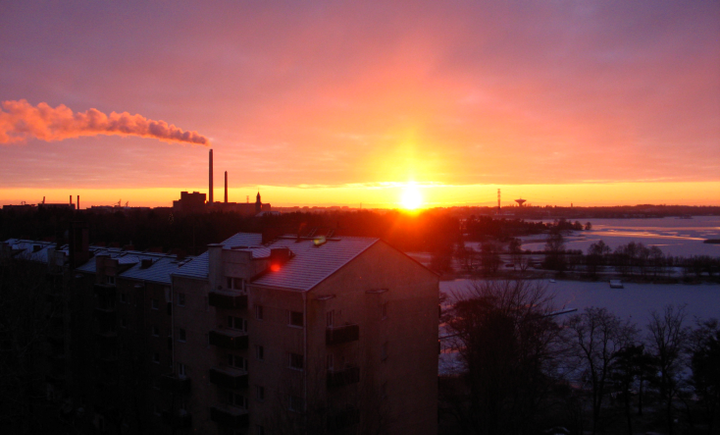 Kaupunkinäkymä, jossa aurinko laskee horisonttiin ja tehtaiden piipuista tulee savua.