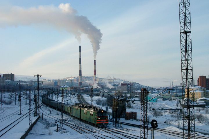 Kuvassa on junaratoja, joilla ajaa yksi juna. Taustalla näkyy tehtaiden piippuja.