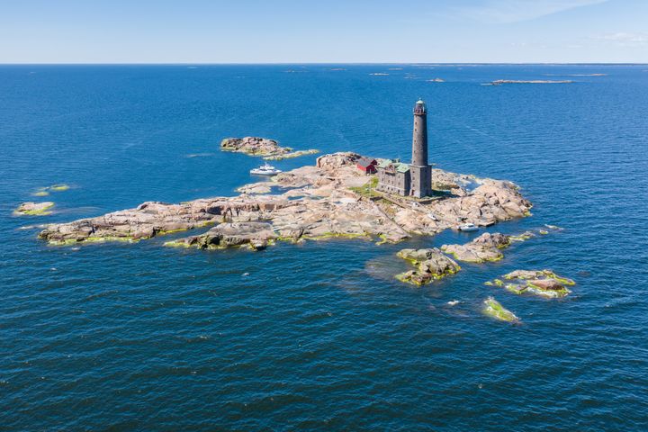 Ilmakuva kallioisesta majakkasaaresta sinisen meren keskellä kirkkaana päivänä. Saaressa on majakka, muutama rakennus ja kaksi venettä rantautuneena kallioiden viereen.