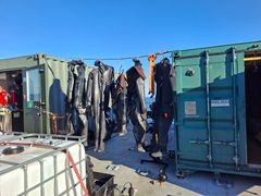 Svarta dykdräkter hänger på galgar för torkning mellan gröna metallcontainrar.