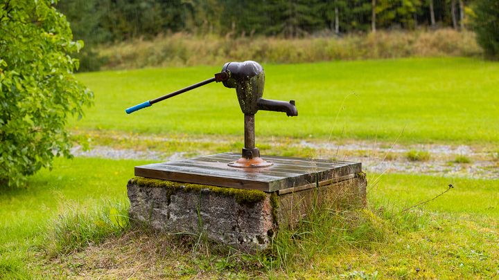 Vanha käsikäyttöinen kaivopumppu kivisen kaivon kannella sateisessa maalaismaisemassa.