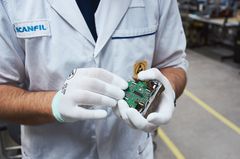 Henkilö valkoisessa työasussa pitelee elektroniikkakomponenttia käsissään.
