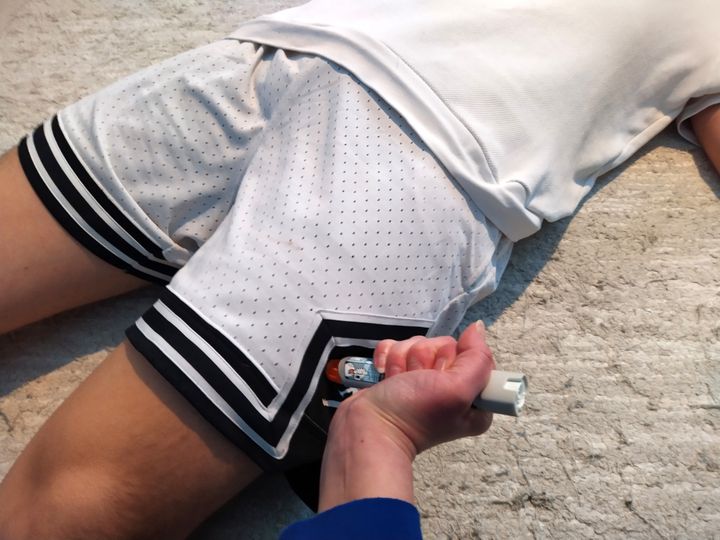 Nuori poika makaa maassa. Aikuisen kädessä adrenaliini-injektori, jolla hän pistää pojan reiteen adrenaliinia.