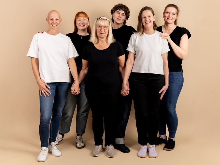 Kuusi eri-ikäistä naista seisoo yhdessä beigeä taustaa vasten.