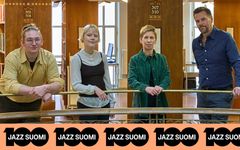 Luotsi-yhtyeen muusikot nojaavat kaiteeseen Kansalliskirjaston sisätiloissa. "Jazz Suomi 100" -logonauha näkyy alareunassa.