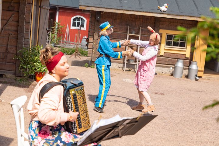 Koiramäen maalaismiljöö punaisine maalaisrakennuksineen ja hiekkapihoineen. Vasemmalla etualalla haitarinsoittaja ja kauempana Koiramäen tanssivia asukkaita.