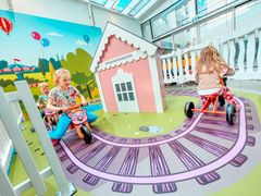 Kolme lasta ajamassa kolmipyörillä leikkipuistossa värikkäiden seinämaalausten edessä.