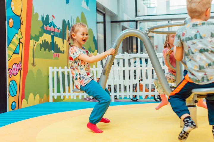 Kolme lasta leikkii värikkäässä sisäleikkipuistossa. Taustalla on piirrosmaisia kuvia ja valkoinen aita.