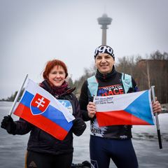 Kaksi henkilöä hymyilee ulkona talvisessa maisemassa, pitäen Slovakian ja Tšekin lippuja. Taustalla näkyy tornirakennus.
