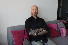 Henkilö istuu kädet ristissä rinnalla sohvalla musta paita päällään, vieressään pinkkejä tyynyjä.