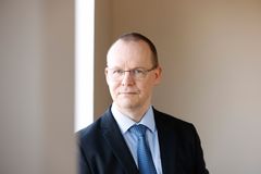 Tummansiniseen pukuun, vaalenasiniseen kravattiin ja valkoiseen paitaan pukeutunut silmälasipäinen mies katsoo lähikuvassa vakavana kameraan.