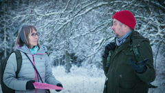Kaksi henkilöä keskustelee talvimaisemassa, lumiset puut taustalla.