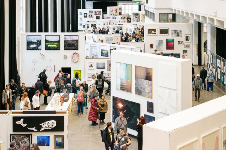 Näyttelytila täynnä ihmisiä katsomassa seinille ripustettuja taideteoksia.