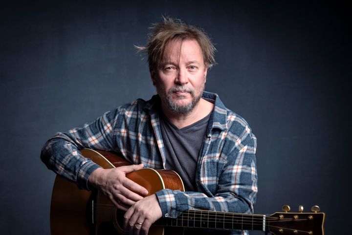 Harmaalla taustalla istuu miesoletettu ruutupaidassaan akustinen kitara sylissään.