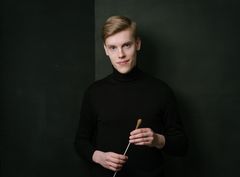 Nuori mies mustassa poolopaidassa pitää kapellimestarin tahtipuikkoa tummalla taustalla.